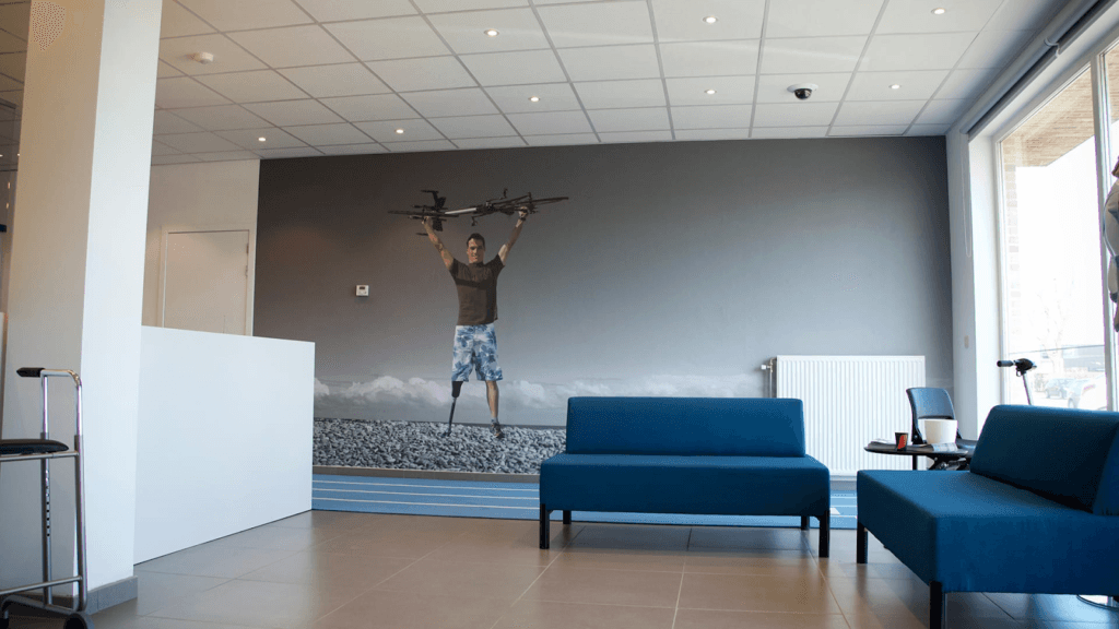 muurfolie met foto van man op een strand in shorts met beenprothese die een fiets boven zijn hoofd houdt, in kantoor met 2 blauwe zitbanken